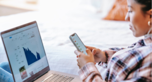 A women is sitting in front of her laptop with he phone in hand. She is looking at housing market analytics.