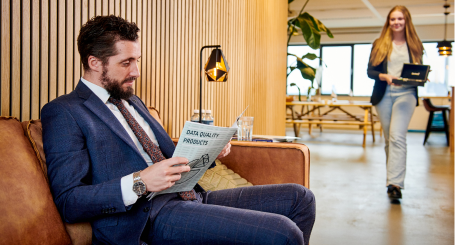 A man in a suit and tie engrossed in reading a magazine while seated on a couch.